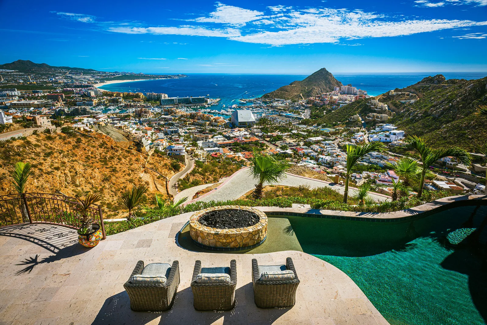 View from a Pedregal hillside terrace with a fire pit and pool, looking over Cabo San Lucas, the marina and Land's End under a blue sky