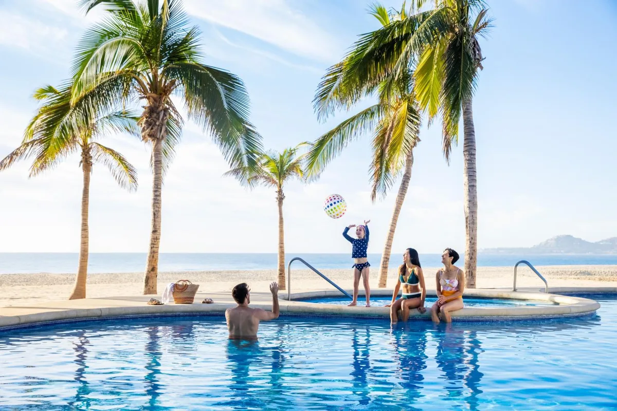 A family plays with a beach ball in the pool at Hyatt Ziva Los Cabos, with palm trees and the ocean behind them