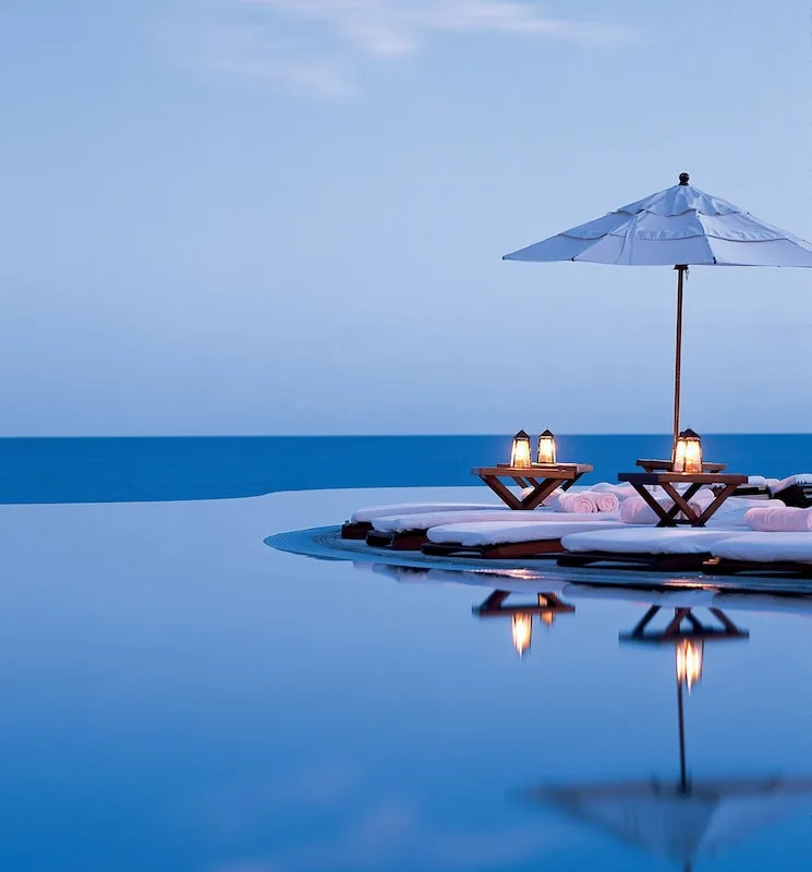 Infinity pool at Las Ventanas al Paraíso at dusk, with an umbrella and candlelit loungers reflected in the water