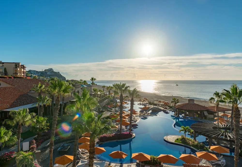 Pools at Pueblo Bonito Sunset Beach with palm trees and orange umbrellas, and the sun shining over the Pacific Ocean behind them