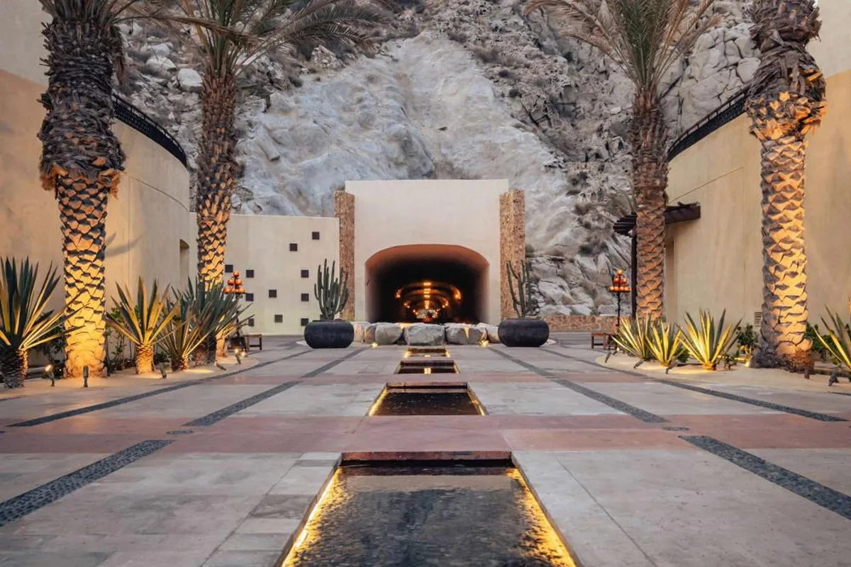 Palm-lined walkway and reflecting pool leading to the lit tunnel entrance of Waldorf Astoria Los Cabos Pedregal, cut into the rock