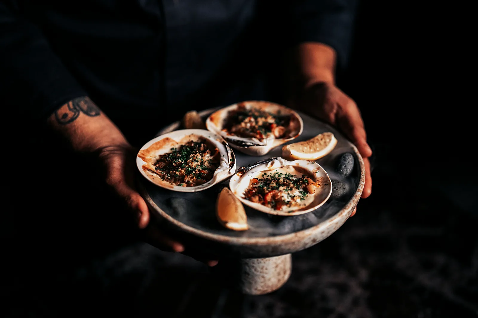 A server holds a stone platter of three baked clams in the shell, topped with herbs and breadcrumbs, with lemon wedges, at HUMO in Zadún