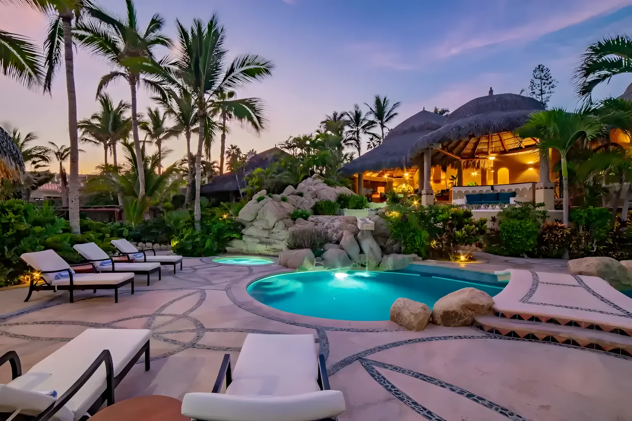 Villa Las Rocas pool at dusk in Costa Brava, lit blue among rock features and palms, with lounge chairs and a palapa roof glowing behind