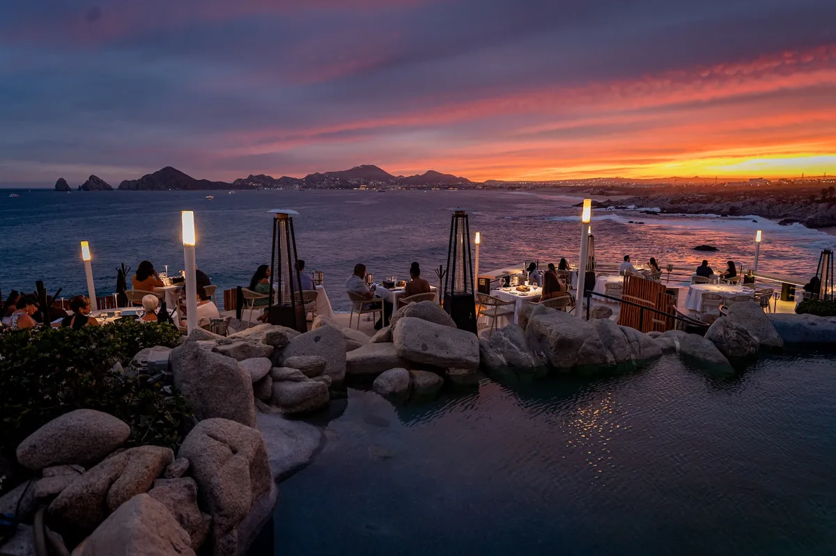 Diners on the cliffside terraces at Sunset Monalisa at dusk, with lanterns lit and the Arch at Land's End on the horizon
